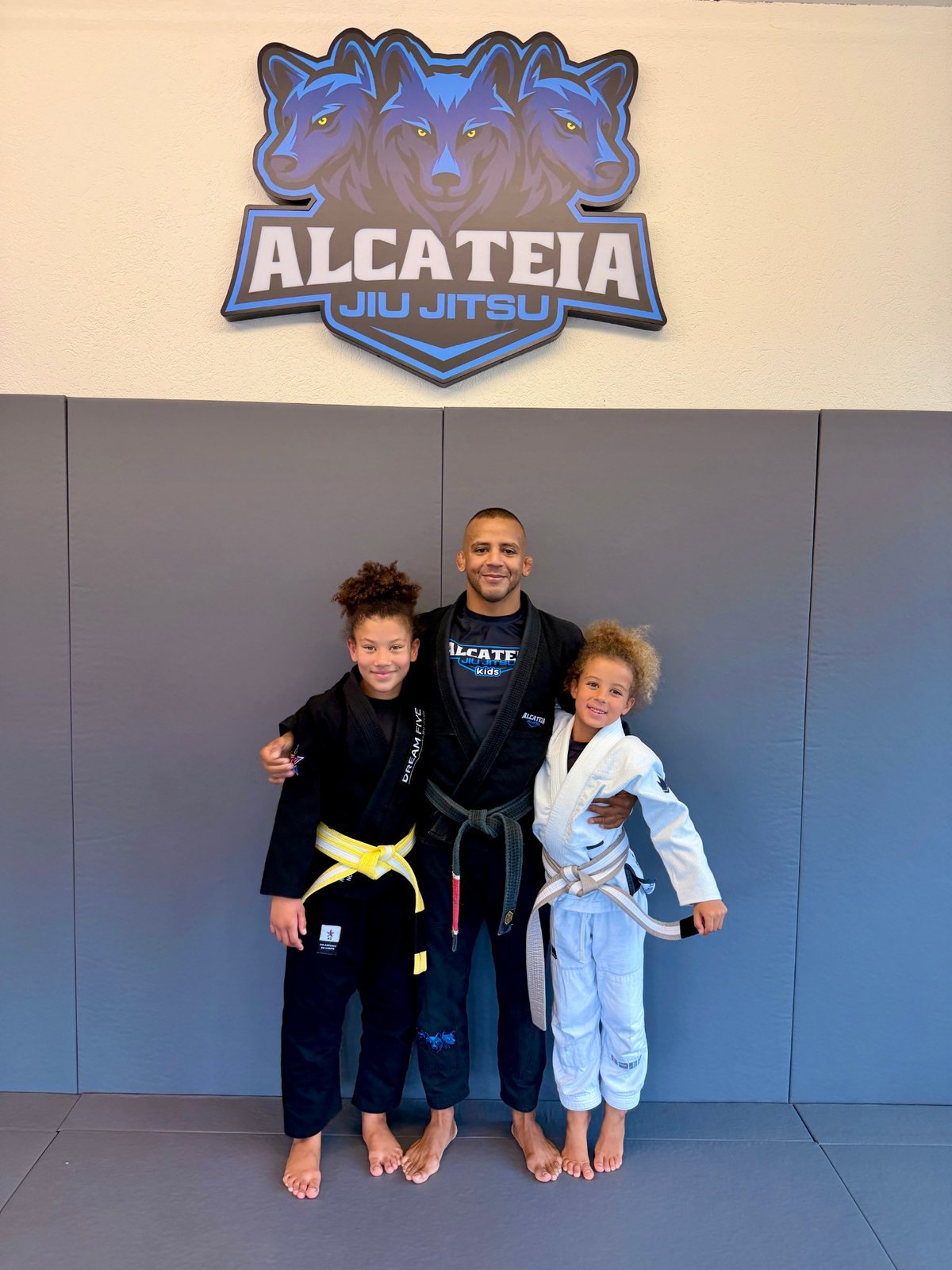 Aleyna in a light-blue gi with her grey-white belt at Alcateia Jiu Jitsu, next to her sister and their instructor.
