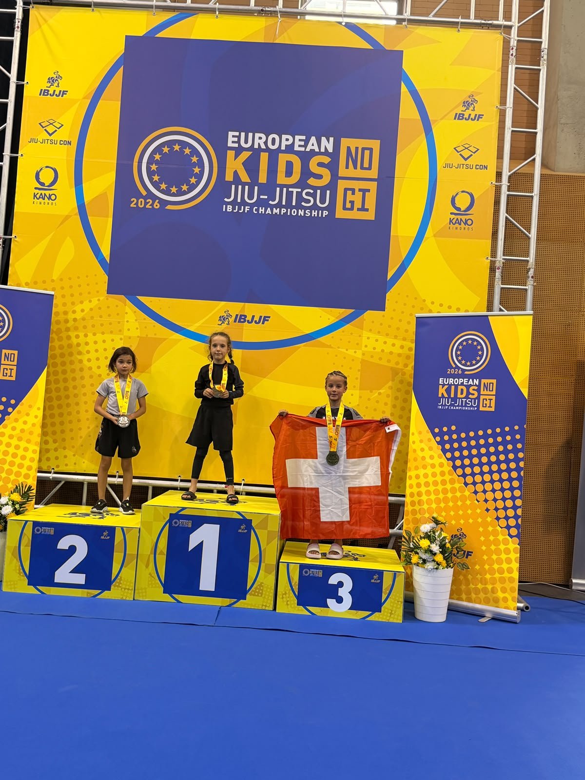 Aleyna on the third step of the podium with the Swiss flag at the European Kids No-Gi IBJJF Championship, Lisbon, October 2026.