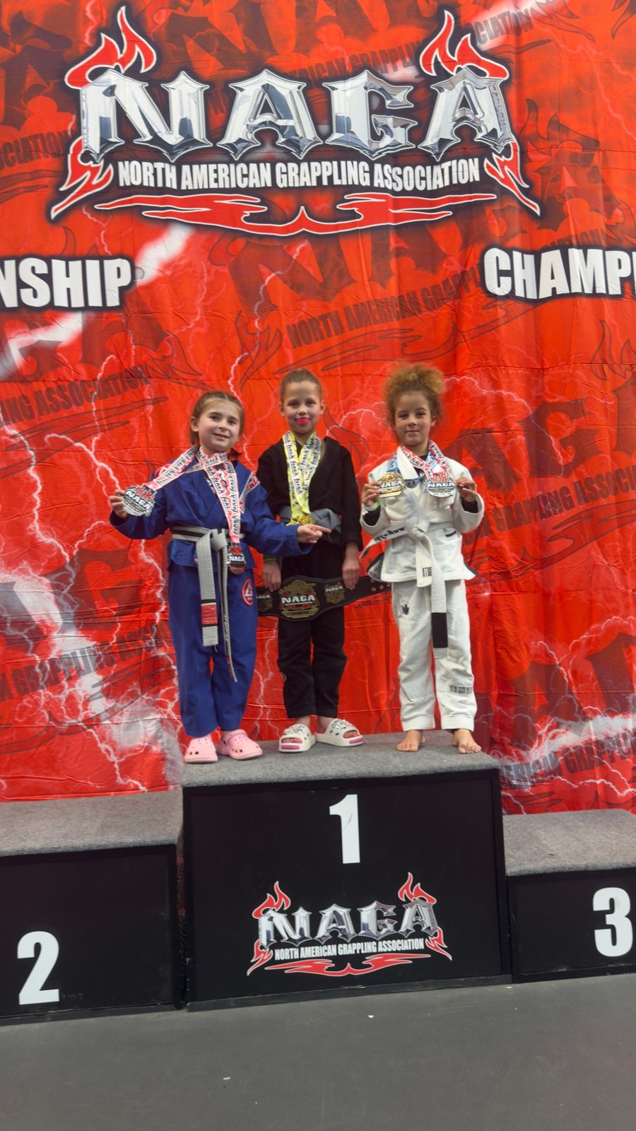 Aleyna in a white gi on the podium at NAGA Paris, March 2026, with her two medals.